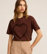 Remera marrón de algodón con cuello redondo y mangas cortas. Presenta un aplique de corazón con volados en el frente.