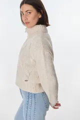 Cardigan color crudo de tejido de punto con diseño trenzado, cuello alto y cierre frontal con cremallera.