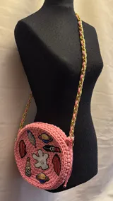 Cartera tejida a crochet color rosa, con forma circular y apliques bordados de labios, plumas, planetas y una mano. Tiene una correa de cadena dorada entrelazada con trapillo rosa, cierre superior con cremallera y forro interior.