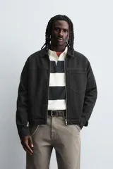 Sobrecamisa de denim de algodón negro con efecto lavado, corte relajado (relaxed fit). Presenta cuello solapa, cierre frontal con cremallera, manga larga con puño abotonado y bolsillo plastrón en el pecho.