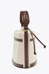 Bolso tipo bucket cilíndrico a rayas verticales en tonos beige y blanco, con asa de mano trenzada color marrón y asa de hombro extraíble. Incluye bolso de tela interior y pespuntes a contraste.