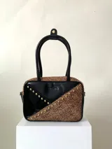 Cartera rígida negra de cuero con tachas plateadas y tres combinaciones de cuero diferentes que aportan textura. Tiene herrajes y cierre metálico plateado marca YKK. Cuenta con un asa fina para llevar al hombro o en la mano y una correa larga extra regulable.