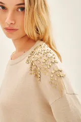 Sweater color beige con cuello redondo y mangas largas. Presenta apliques dorados en los hombros y un cinturón marrón con forma de flor.