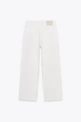 Pantalón cropped color beige de tiro alto y corte recto, confeccionado en algodón. Cuenta con bolsillos delanteros y traseros, trabillas en la cintura y cierre frontal con cremallera y botón.