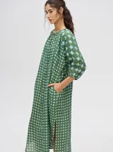 Vestido largo con mangas 3/4 y cuello mao, confeccionado en tela con estampado de círculos en tonos verde oliva y celeste pálido. Presenta cierre frontal de botones marrones y corte amplio con aberturas laterales.