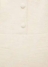 Mono largo color crudo confeccionado en tejido tweed, con cuello redondo, sin mangas y cierre frontal con botones. Presenta dos bolsillos de ribete delanteros y diseño recto.