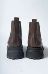 Bota corta tipo Chelsea de cuero negro, con puntera cuadrada y suela track gruesa y dentada. Presenta paneles elásticos acanalados a los lados para facilitar el calce.