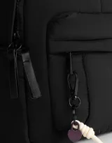 Mochila color crema de tela acolchada con pespuntes verticales. Tiene un bolsillo frontal con cierre, doble cierre superior, manija superior, correas regulables y bolsillos laterales. Incluye un charm colgante de corazón.