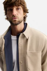 Sobrecamisa negra de tejido con elasticidad, cuello solapa y manga larga acabada en puño con botón a presión. Tiene bolsillos de plastrón en el pecho y cierre frontal con botones a presión.