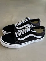 Championes Vans UA Old Skool, color negro con la icónica franja lateral Sidestripe™ blanca. Presentan una silueta baja con cordones blancos, parte superior de gamuza y lona resistentes, y cuello acolchado. La suela es de goma con patrón waffle.