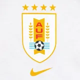 Remera Nike blanca de manga corta con cuello redondo. Presenta el escudo de la Asociación Uruguaya de Fútbol (AUF) en dorado y negro, con cuatro estrellas doradas encima, y el logo de Nike dorado debajo.
