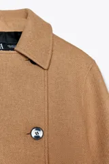 Abrigo color camel confeccionado en hilatura con lana. Tiene cuello solapa, manga larga, bolsillos delanteros ocultos en costura y cierre frontal cruzado con botones negros.