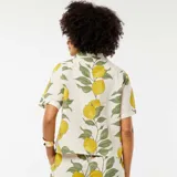 Blusa color crema con estampado de limones y hojas verdes, de manga corta, cuello camisero y cierre frontal con botones.