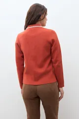 Chaqueta de tejido color terracota/lacre, con cuello y bordes de bolsillo en blanco con textura de punto. Posee cierre frontal de botones y manga larga.