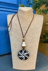 Collar con cordón marrón y dije circular de resina negra con diseño de rayos blancos. Incluye cuentas de madera clara y oscura.