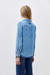 Camisa de denim celeste con cuello clásico, cierre frontal con botones a presión, dos bolsillos con solapa en el pecho y mangas largas con puños abotonados.