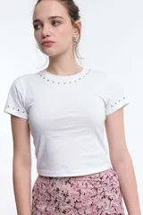 Remera blanca de algodón con cuello redondo y mangas cortas. Presenta pequeños apliques metálicos plateados alrededor del cuello y en las mangas.