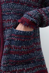 Chaqueta corta de tejido bouclé o tweed, sin cuello ni cierre frontal, con ribetes en color bordó. El tejido es una mezcla de hilos en tonos azul marino, rojo/bordó y blanco, creando un efecto jaspeado. Los puños tienen un detalle de flecos en color bordó.