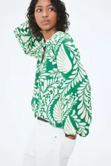 Blusa de tejido liviano color verde con estampado de hojas blancas, cuello corto fruncido y tapeta de botones. Mangas globo largas con volumen de corte raglán y puños finos abotonados.