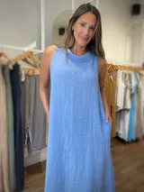 Vestido largo sin mangas color azul claro, confeccionado en lino, con cuello tipo buche o drapeado. Presenta un calce holgado y bolsillos laterales.