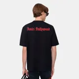 Camiseta negra de corte regular con cuello redondo y manga corta. Presenta un estampado frontal en color rojo/rosado con el texto "Amiri Hollywood" en tipografía gótica y una estrella estilizada con las letras "M" y "A" debajo.