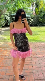 Vestido corto confeccionado en tela de lentejuelas negras, con escote recto y breteles finos. Presenta detalles de flecos de rafia en color rosado tanto en el escote como en el ruedo.