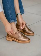 Zapato acordonado color bronce de cuero vacuno con perforaciones decorativas, taco de madera con folia en cuero de 4cm y entre suela de goma de 1,5cm. Horma de punta redonda y cómoda.