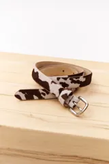 Cinturón de cuero con estampado animal print de vaca en tonos marrón y blanco, con hebilla metálica plateada.