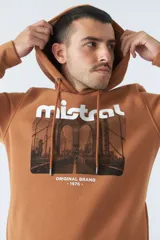 Buzo marrón con capucha y bolsillo canguro. Presenta un estampado frontal con el logo de la marca Mistral y una imagen de un arco arquitectónico. Confeccionado en algodón y poliéster.