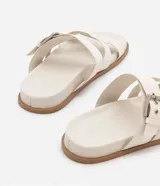 Sandalias planas de cuero sintético color blanco, con puntera redonda y dos tiras, una con hebilla metálica y otra en forma de H.