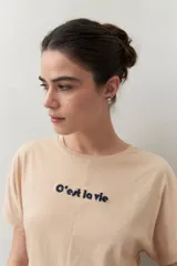 Remera blanca de manga corta con bordado frontal "C’est la vie", calce relajado y frunces laterales.