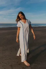 Vestido largo blanco de lino con corte recto, cuello en V y mangas cortas. Presenta ribetes azules en cuello, mangas y laterales. Tiene aberturas laterales en el ruedo.