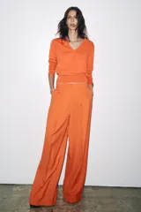 Pantalón ancho color naranja, de tiro medio y cintura con trabillas. Tiene bolsillos laterales y de vivo en espalda, pinzas en delantero, raya planchada y cierre frontal con cremallera, botón y ganchos metálicos.