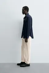 Pantalón chino negro de corte relajado (relaxed fit) con tiro medio, confeccionado en algodón con elasticidad. Presenta bolsillos delanteros y bolsillos traseros de vivo, con cierre frontal de cremallera y botón.