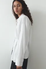 Camisa blanca de cuello solapa y manga larga. Detalle de pliegues con botones. Cierre frontal con botones.
