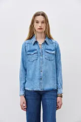 Camisa de denim celeste con cuello clásico, cierre frontal con botones a presión, dos bolsillos con solapa en el pecho y mangas largas con puños abotonados.