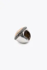 Maxi anillo metálico plateado con forma ovalada y pieza incrustada labrada dorada a contraste. Detalle de aplicación de brillos negros alrededor de la pieza dorada.