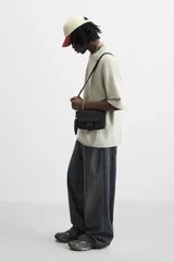 Bandolera negra de tela técnica con correa ajustable y extraíble. Tiene un compartimento principal con cierre de cremallera, un bolsillo interior con cremallera, un bolsillo frontal con solapa y hebilla de liberación rápida, y un monedero removible.