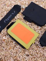 Tarjetero Rusty Runoi con cierre y correa para colgar, confeccionado en poliuretano color verde con aplique naranja y logo de la marca.