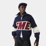 Chaqueta de tejido punto grueso color azul marino con franjas horizontales en color crema y un gran gráfico tejido en color bordó con borde crema en el pecho. Presenta cuello alto y cierre frontal metálico.