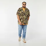 Camisa de manga corta con estampado de hojas tropicales en tonos verdes y marrones sobre fondo negro. Tiene cuello clásico, corte recto y cierre frontal con botones.