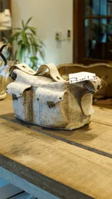 Bolso con textura craquelada en tonos beige y marrón claro, con doble asa corta, tres bolsillos frontales con cierre de broche metálico y detalle de ojales metálicos en la parte superior.