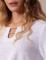 Blusa blanca de tejido labrado con cuello redondo, escote en V y aplique metálico.
