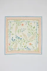 Pañuelo cuadrado de tela color beige claro con estampado botánico de flores y hojas en tonos pastel (azul, salmón, verde y amarillo). Presenta un borde decorativo con un patrón de espirales en color salmón y un borde exterior azul claro.