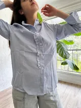 Camisa celeste y blanca a rayas verticales, con cuello con volados, cierre frontal con botones y mangas largas con puños abotonados.