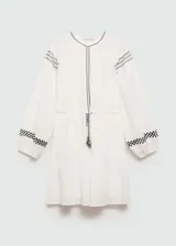 Vestido corto blanco de algodón con bordados y detalles calados. Tiene cuello redondo, mangas largas con puños elásticos, cintura elástica con lazo y diseño evasé.