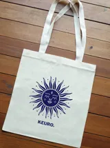 Bolso de mano tipo tote bag color crudo, con asas de tela y estampado de un sol con rostro y la palabra "KEURO." en azul.