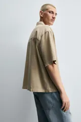 Camisa de denim de algodón, color beige claro, con corte relajado, cuello bowling y manga corta. Presenta cierre frontal con botones a presión y un pequeño bordado a tono en el pecho.