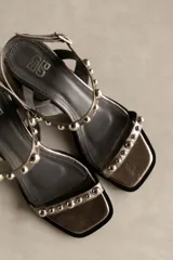 Sandalia de cuero color marrón con taco bloque medio y punta cuadrada. Presenta tachas esféricas doradas en las tiras y hebilla de ajuste en el tobillo.