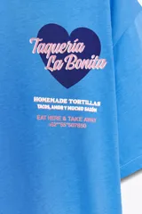 Camiseta celeste de algodón con corte oversize, cuello redondo, manga corta con hombro caído y estampado frontal con texto 'Taquería La Bonita' y un corazón.